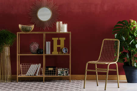 Gold chair and plant near shelves against red wall with round mirror in elegant living room interiorの写真素材