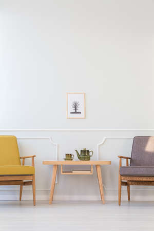 Wooden table between grey and yellow armchair against white wall with poster in living room interior