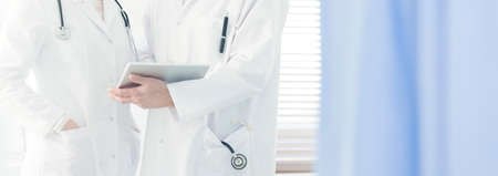 Close-up of a doctor in white coat with stethoscope holding a tablet, consulting on a case with a colleague in bright hospitalの写真素材
