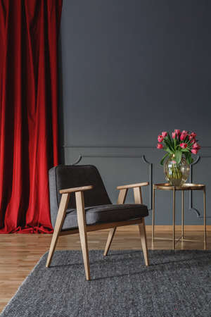 Grey wooden armchair on carpet next to gold table in living room interior with red drapesの写真素材