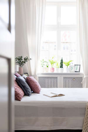 Book and pink pillow on bed in white bedroom interior with window. Real photo