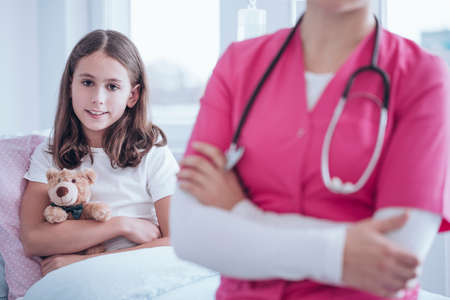Smiling girl with plush toy during stay in the clinic. Blurred caregiver wearing pink uniform in the foregroundの写真素材