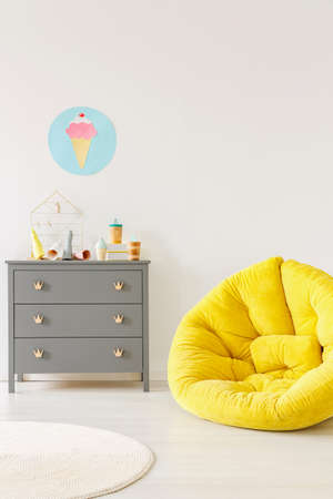 Yellow pouf next to a grey chest of drawers and ice-cream poster on a white wall in kid room...