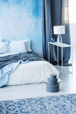 Patterned carpet and grey boxes next to bed with blue bedding in minimal bedroom interior with lampの写真素材