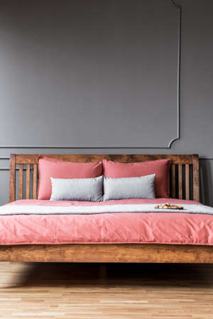 Close-up of a simple bed with wooden bedhead and pink sheets in dark, elegant bedroom interior with molding on dark walls. Real photoの写真素材