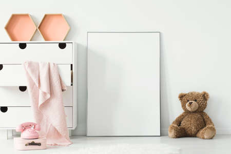 Teddy bear next to white empty poster in baby's room interior with pink blanket in cabinet. Real photo. Paste your graphic hereの写真素材