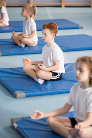 Elementary school pupil and other children sitting on mats with their legs crossed and eyes closed in relaxing meditation classの写真素材