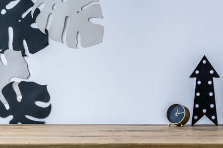 Gold clock and black arrow on wooden desk against white wall with monstera leaves in workspace interiorの写真素材