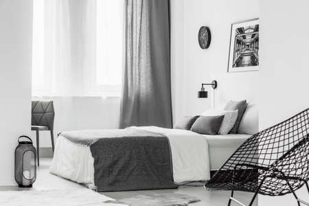 Lantern next to bed with blanket in bright grey bedroom interior with armchair and curtainの写真素材