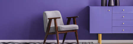 Grey armchair standing next to a purple cupboard in violet room interior with place for your table