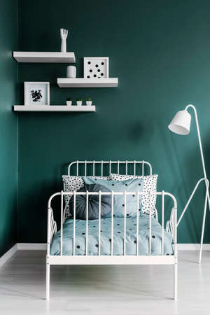 Shelves with decorations and a floor lamp above a white, metal frame twin bed in a dark green room...