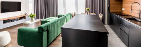 Side angle of a spacious kitchen interior with dark cupboards, kitchen island, corner sofa and tv