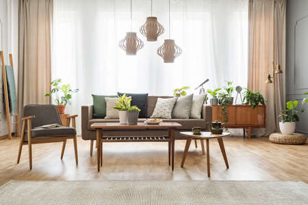 Grey armchair next to wooden table and sofa with cushions in flat interior with drapes and lamps....