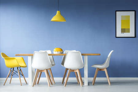Minimal, modern dining room in interior with a table and contrasting white and yellow chairs around it, against blue wall with a mockup posterの写真素材
