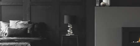 Real photo of an elegant lamp with a glass base on a simple night stand between a cozy bed and fireplace in black apartment room interiorの写真素材