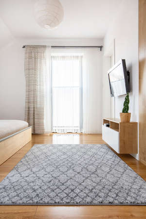 Grey carpet on wooden floor in bright bedroom interior with window and television. Real photo