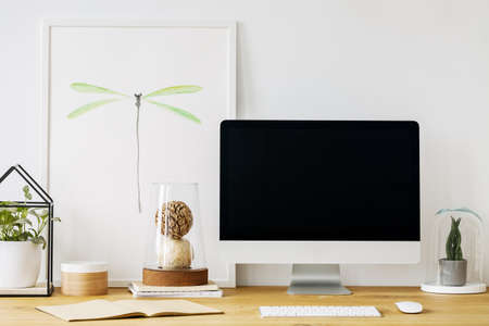 Real photo of a desk with an empty computer screen, ornaments and a poster in a room interiorの写真素材