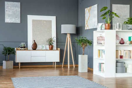 White cabinet and shelves, wooden lamp and paintings on the grey wall in living room interiorの写真素材
