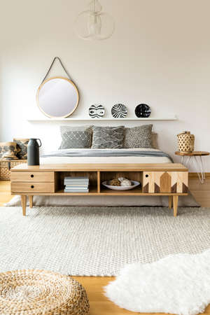 Round mirror and patterned plates above bed in scandi bedroom interior with wooden rustic cupboard