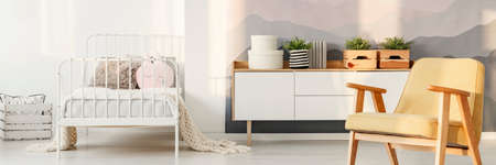 Yellow wooden armchair and plants on white cupboard in cozy bedroom interior with metal bed