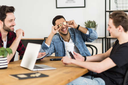 Friends having fun at the meeting while one is putting bitcoins to his eyes