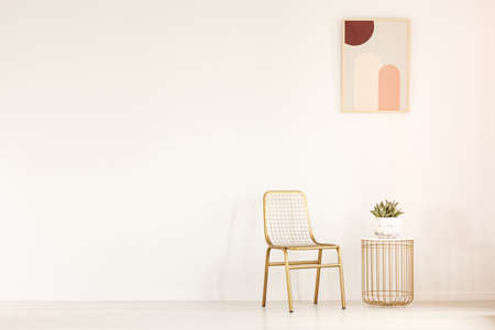 Real photo of a gold chair standing next to a small table with a plant in minimal living room...