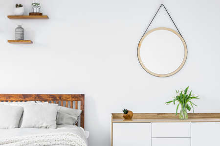 Tulips on a scandinavian style dresser, wooden shelves with plants and a vase and round mirror in a white bedroom interiorの写真素材