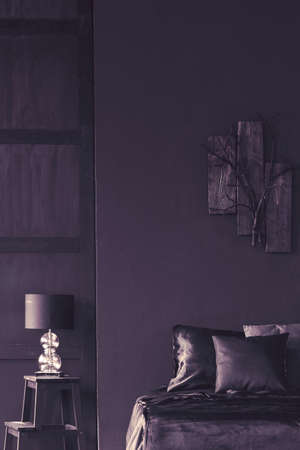 Lamp on a stool next to bed in dark violet bedroom interior with wooden decor on the wallの写真素材