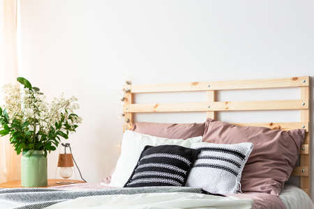 Real photo of a bed with cushions and a wooden bedhead standing next to a table with a plant in simple bedroom interiorの写真素材