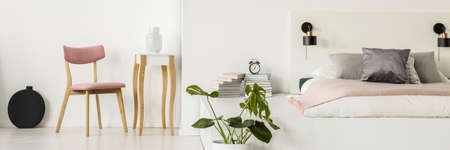 Pink chair standing by small table with vase in white bedroom interior with fresh green plant, clock...