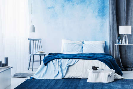 Navy blue carpet in minimal bedroom interior with blanket on bed next to a chair and under a lampの写真素材