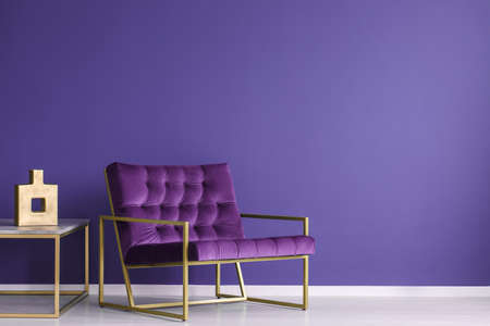 Purple armchair next to a table with gold vase in elegant living room interior with copy space on the wallの写真素材