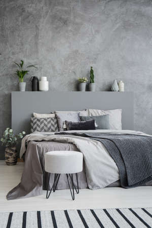 Striped carpet and stool in grey bedroom interior with sheets on bed with headboard. Real photoの写真素材