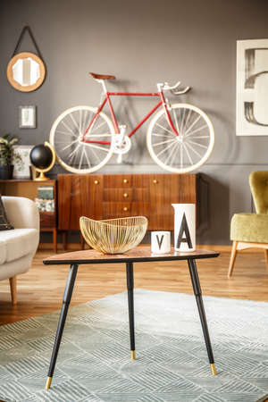 Close view of a black small table with a fruit bowl standing on a green rug with a red bicycle on a sideboard in the background against gray wall in a vintage interior. Real photo.の写真素材
