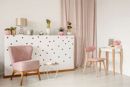 Elegant armchair and an open book by a polka dot wall, wooden vanity table and curtains in a white bedroom interior with dirty pink elementsの写真素材