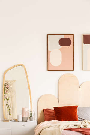 Real photo of a bed with red and grey cushions standing next to a cupboard with a mirror and a wall with two posters in rusty bedroom interiorの写真素材