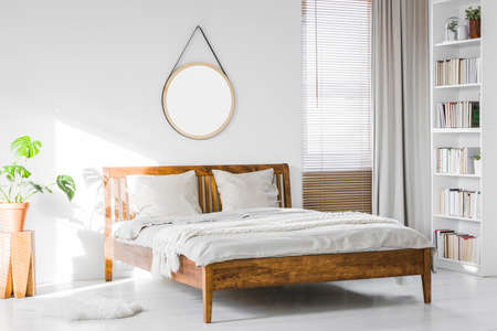 A wooden rustic bed frame and a home library bookcase in a natural, sunlit hotel room interior with white wallsの写真素材