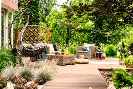 Single swing and garden furniture set on a wooden terrace in a green garden with trees and bushesの写真素材