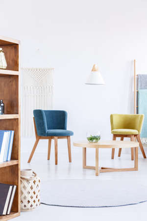 Real photo of a blue and yellow armchair standing next to a wooden table in bright white living room...