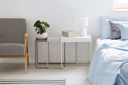 Plant, lamp and books on table between armchair and blue bed in simple bedroom interior. Real photo