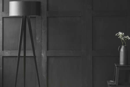 Black lamp near white flowers on stool in unicolor living room interior. Real photo