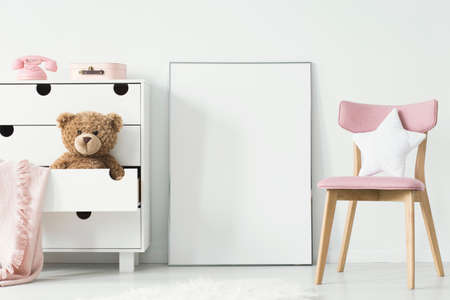 Empty poster with place for your graphic standing on the floor in white room interior with pink wooden chair and teddy bear placed in cupboard's drawerの写真素材