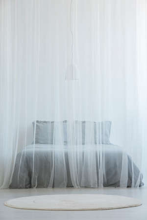 Mosquito net bed with grey bedding standing in white room interior with round rug on the floorの写真素材