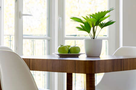 Plant and apples on wooden table in white dining room interior with window. Real photoの写真素材