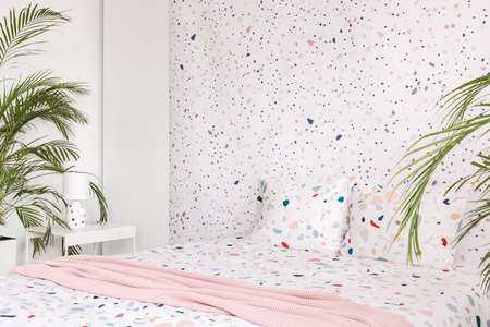 Pink blanket on patterned bed with pillows in bright bedroom interior with plants. Real photo