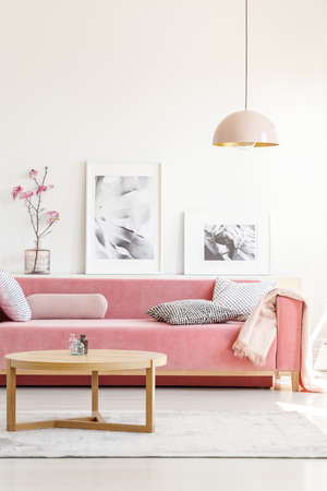 Real photo of a pink sofa with pillows standing behind a table, in front of a shelf with posters and...