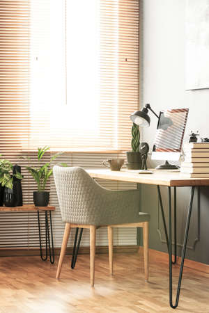 Grey armchair at desk with desktop computer and lamp in bright workspace interior. Real photoの写真素材