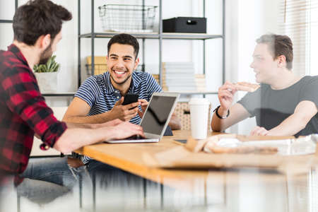 Smiling man using smartphone while his friends eat pizza and use a laptop