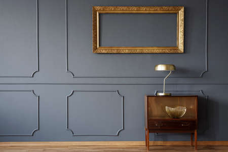 Vintage wooden cabinet and an elegant, empty golden frame on a dark wall with molding in a luxurious living room interiorの写真素材