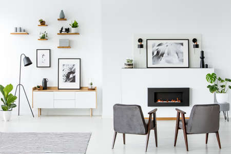 Grey armchairs in front of fireplace under poster in white apartment interior. Real photoの写真素材